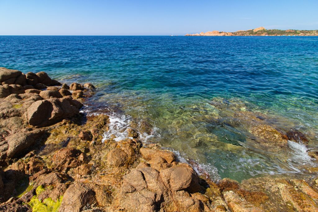 A landscape shot of big rocks, green hills in a blue ocean with a clear blue sky